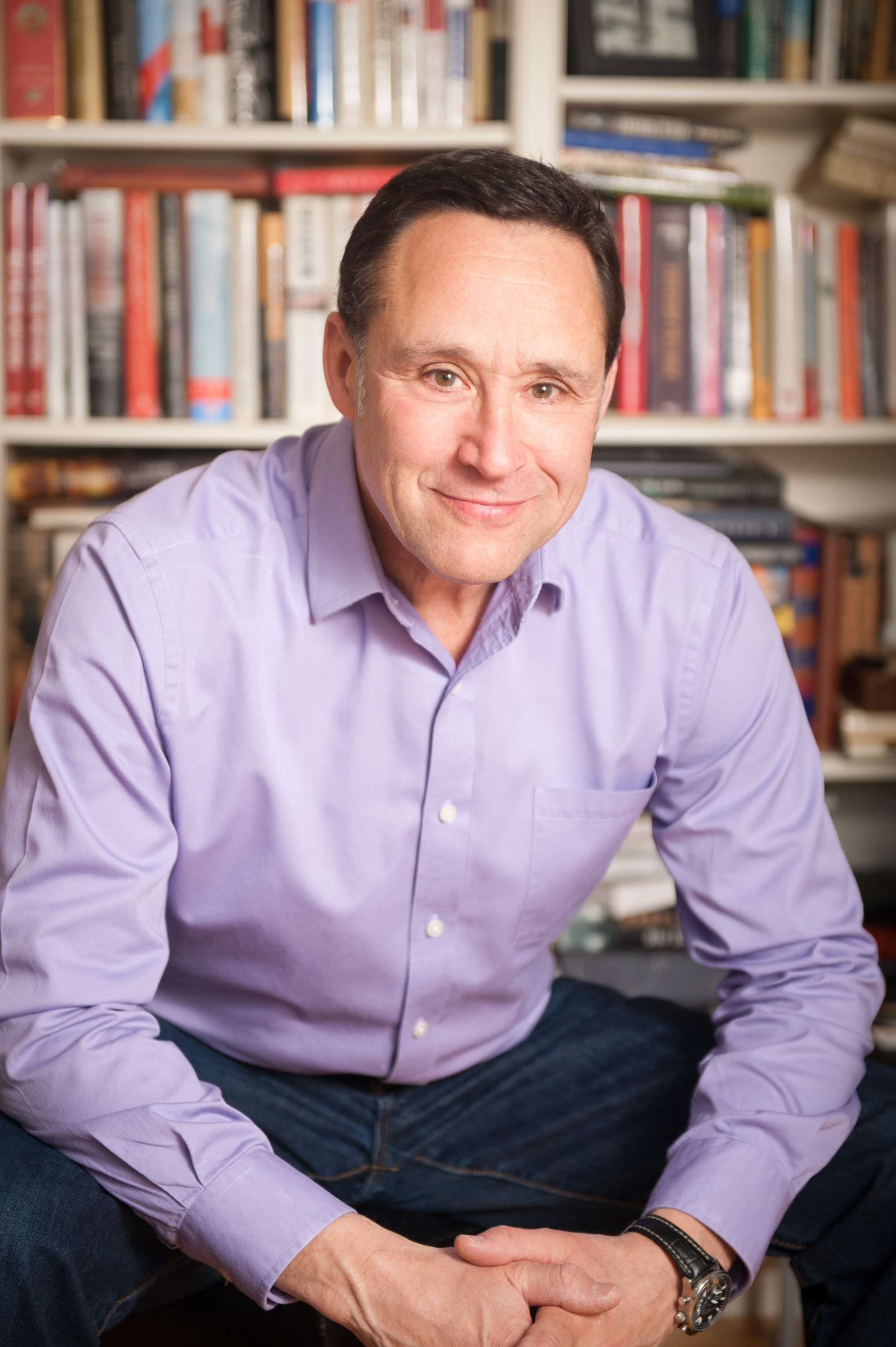 Professional photograph of author Ted Reinstein in a crisp lilac button-down posed sitting in front of a full bookcase