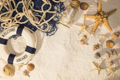 A life preserver, navy and tan rope, seashells, starfish, and a glass bottle filled with seashells laying atop a light sandy backdrop