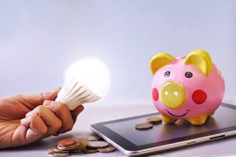 A cute piggy bank standing atop an ipad with coins splayed about while an outstretched hand holds a lightbulb