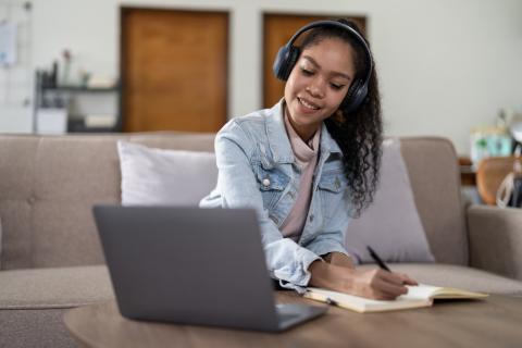 Young woman sits on a couch at home with headphones on in front of her laptop, taking notes in a small journal