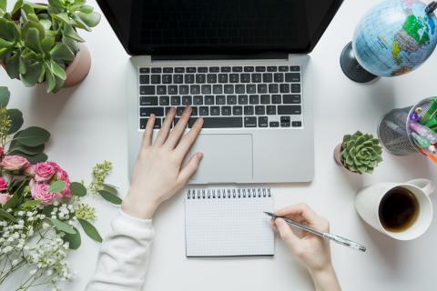 Aerial view of a laptop surrounded by plants, a globe, and writing utensils with a hand on the keyboard and another hand taking notes