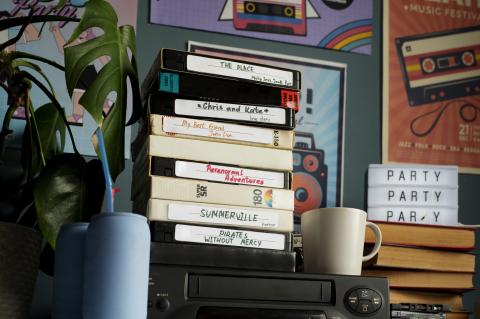 Stack of VHS tapes with labels written in a variety of colorful handwritten scrawls sitting atop a VCR next to a potted fern and a cream-colored mug 