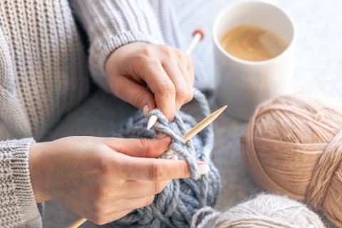 Close up photograph of a woman's hands knitting with a pale blue yarn next to a mug of coffee