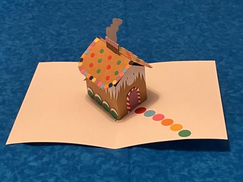 Photograph of a three-dimensional paper-cutout facsimile of a gingerbread house propped atop a sepia sheet of paper with a polka dot pathway to its "entrance"