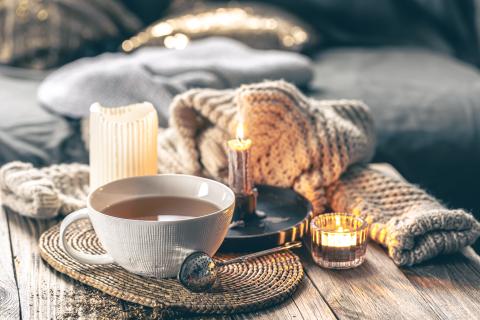 A photo with a cozy atmosphere and warm lighting, depicting a small teacup of tea with milk, a thick wool knitted scarf, and candles on a wooden table in front of a slate blue sofa