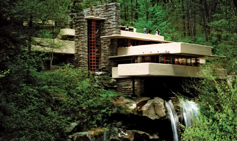 Photograph of Frank Lloyd Wright's "Fallingwater" (1935) landmark architectural design in Mill Run, Pennsylvania - made up of a series of stark rectangular prisms split between mahogany colored scaffolding and stonework pillars, all of which is nestled into a dense forest foliage and stood atop naturalistic stonework and a perpetual waterfall