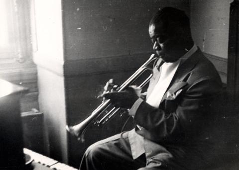 Black-and-white photograph of Louis Armstrong playing the trumpet while sitting before a grand piano, light streaming in from a corner window to a chiaroscuro effect