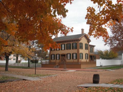 Photograph of Abraham Lincoln's family home: a subdued yellow ochre two-story New England -style corner-street home with brown trim and dark window shutters, vibrant autumn trees in the foreground bookending visual of the home