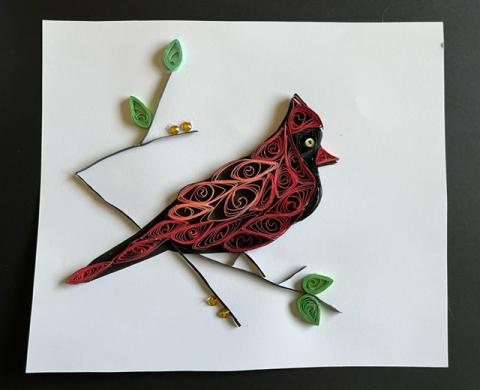 Photograph of a stylized craft-work emulation of a red male cardinal stood atop a narrow branch, the totality of its shape made up of spiraling coils of colored paper