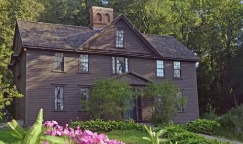 Photograph of "Orchard House," the Alcott family home in Concord, MA, a deep grey-brown home nestled into a thick deciduous wood, the leaves a vibrant springtime green, fluorescent magenta-pink flowers peaking in the foreground