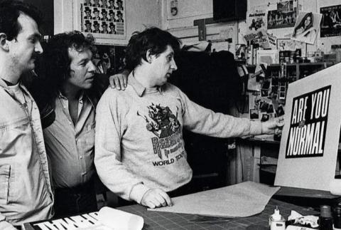 Black and white photograph of Peter Christopherson, Aubrey Powell and Storm Thorgerson of "Hipgnosis" design group in their studio collectively examining their work - an album over with stark sans-serif white text over a black backdrop reading "ARE YOU NORMAL."