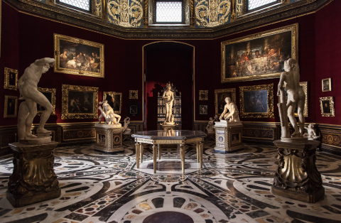 Photograph of the interior to the Uffizi Gallery in Florence, Italy, featuring an ornate marble floor with interlocking, abstract circular shapes of starkly contrasting hues, atop which stand pale marble statues on pedestals contorted in various stages of action, while along deep crimson walls hang Renaissance-era paintings in thick gilded frames, shone brightly in the dim room with spot-lighting