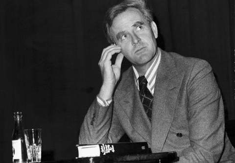 Black and white photograph of author and ex-agent of British Intelligence John le Carré, poised against a stark black backdrop, sitting with one elbow propped against a desk, looking inquisitively out of frame