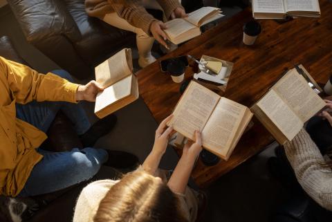 Top-down photograph of a group of people with open books in their hands sitting around a wooden table atop which sits a couple to-go paper coffee cups and a slice of cheesecake