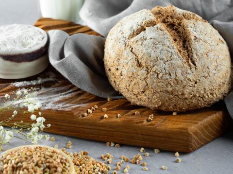 Photograph of a loaf of Irish Soda Bread with a cross-shaped slice down the center, nestled into a light slate-blue blanket, sitting atop a wooden cutting board with a bowl of buckwheat to the side