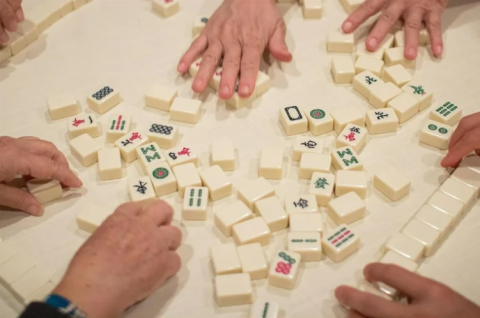 Four pairs of hands working over a set of off-white Mah-Jongg tiles on a matte white table