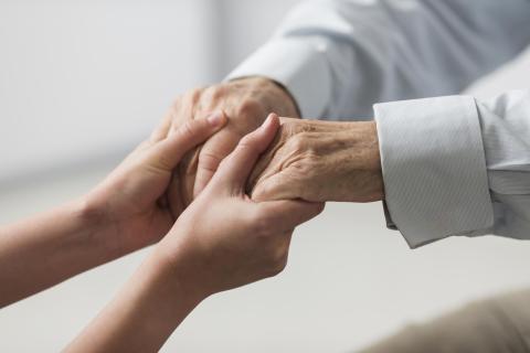 Photograph of two pairs of hands intertwined, one older, the other younger, standing before a light backdrop with soft light diffusing in