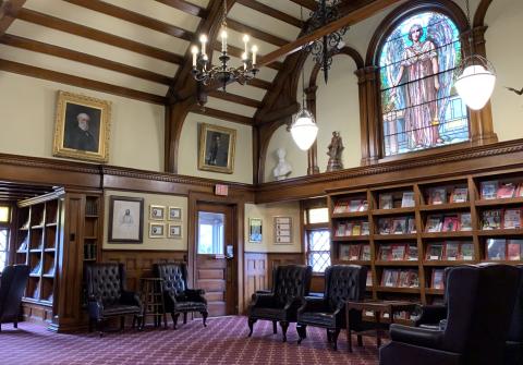 Interior photograph of the historic area of the Emma S. Clark Memorial Library, currently used as its Periodical Room, boasting deep warm wood accents spread across light sepia walls, deep magenta leather chairs, a chandelier, and a stained glass window featuring the archangel Michael.