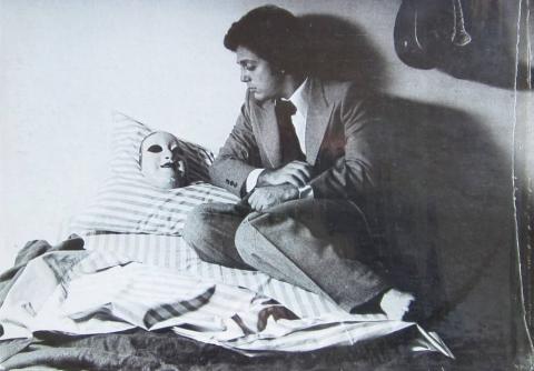 Cropped image from the cover to Billy Joel's 1977 album "The Stranger," in which he singer sits casually on a bed atop a wrinkled blanket, facing an eerie white mask on the pillow beside him.