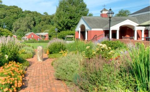 Vibrant daytime photograph of the Long Island Museum, blue skies and full trees in the background, tall foliage and flowers in the foreground, and the red-and-white building nestled within the plantlife.