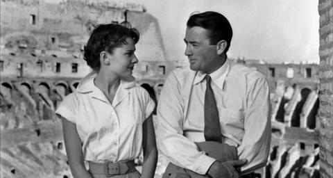 Screenshot from 1953's "Roman Holiday," shot in black-and-white, with lead actors Audrey Hepburn and Gregory Peck sitting in central frame facing one another with the historic Roman landscape in the backdrop.