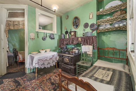 Photograph of an interior space of the Tenement Museum in NYC, showcasing a historically-decorated room with an old-fashioned stove, a simple wooden bedframe, an archival newspaper, among other items intrinsic to the immigrant lives in the 19th century. 