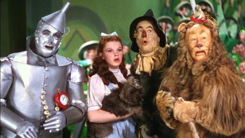 Screenshot from the 1939 film "The Wizard of Oz," featuring the central cast members (the Tin Man, Dorothy, the Scarecrow, and the Lion) faced forward with concerned expressions before a blue-green backdrop.