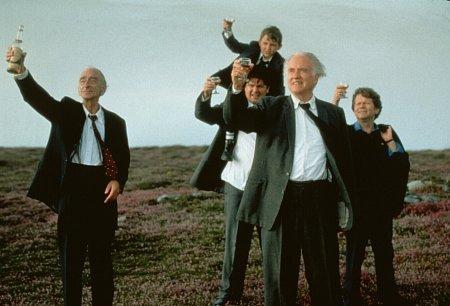 Screenshot from the 1998 film "Waking Ned Devine" featuring five central characters in what appears to be mourning attire, raising glasses on a green hillside to toast a fondly remembered friend.