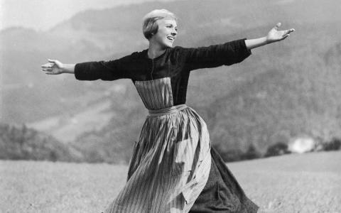Black-and-white screenshot from "The Sound of Music" starring Julie Andrews, featuring the lead actress with her arms spread wide as she sings atop a hill, her skirt spiraling around her as she turns.