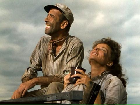Screenshot from the 1951 movie "The African Queen" featuring stars Humphrey Bogart and Katharine Hepburn looking haggard but happy, looking off into the distance before a cloudy slate-colored sky.