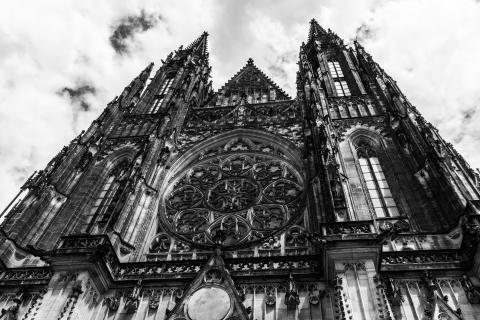 Black-and-white photograph of a gothic-style cathedral, with a series of dark and intricate designs making up the angular silhouette of the tower as viewed from below against a bright but cloudy sky.