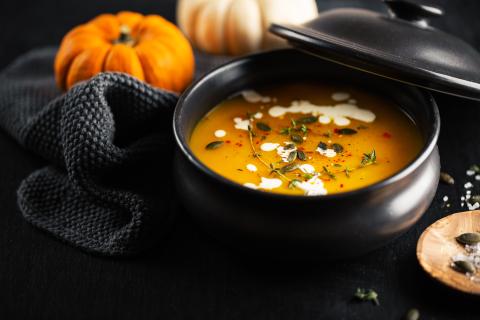 Photograph of a dark kitchen table atop which sits a ramekin filled with a vibrant orange pumpkin soup sprinkled with parsley and heavy cream dollops, nestled into a dark tablecloth amongst which sits whole pumpkins (one white, one orange)