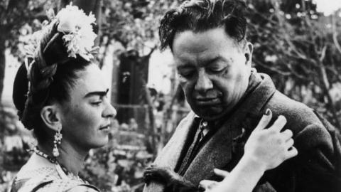 Black-and-white photograph of famed creative couple Frida Kahlo and Diego Rivera, with the former gently caressing the front suit of her partner as he looks down at her extended hand