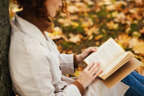 Photograph of a woman in an off-white trench and umber turtleneck sitting leaning against a tree with a book in hand, surrounded by autumn leaves