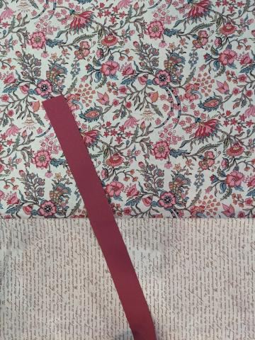 Photograph of a paper-craft bookmark made up of two uniquely patterned papers (a pink-and-green ornate floral, as well as a sepia sheet covered in brown illegible cursive text), with a red-umber ribbon laid across