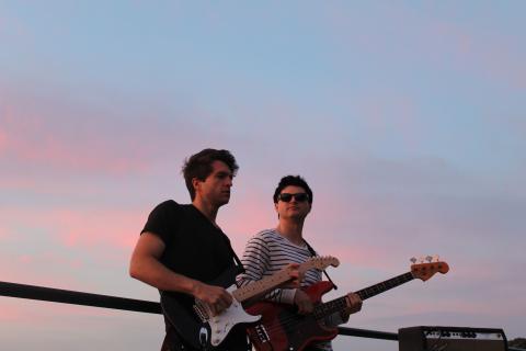 Photograph of the titular members of the Como Brothers band, the two young men viewed from a low angle in dim lighting before a blue-and-pink evening sky