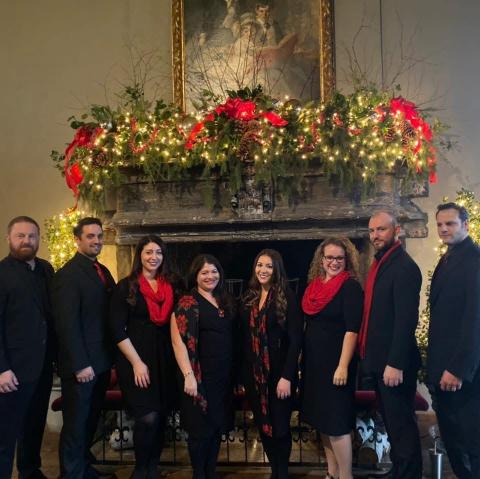 Photograph of the members of the "House of the Red Hart" band, a grouping of eight members wearing all black with red accents, stood in front of a massive fireplace decorated with green faux-foliage, red ribbons, and twinkling golden tealights
