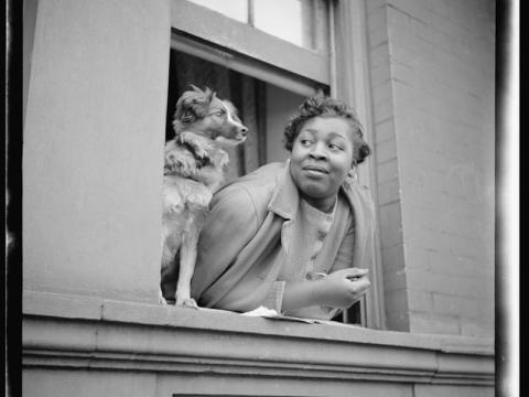 Black-and-white photograph from 1943, depicting a woman leaning out of her Harlem brownstone window with her dog perched just beside her, looking outward together
