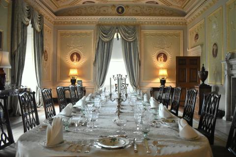 Photograph of a glamorous historic dining room with a long table, dining cloth, decorative napkin displays atop ceramic plates, while in the backdrop there is an ornate mint-and-white wall ornamentation and matching drapery, wooden chairs of a deep brown, and warm dim lighting