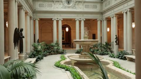 Photograph of the Frick Museum's central "Oasis Room," featuring a series of marble columns enclosing an oblong rounded chamber, at the center of which sits a fountain, above which is a grid-bounded glass ceiling, and along the sides of which stand lively green plants and dark marble statuettes