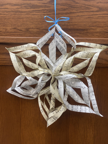 Photograph of a 3D craft snowflake made of cut strips of sheet music and tied up with a blue ribbon and propped on a woodgrain table
