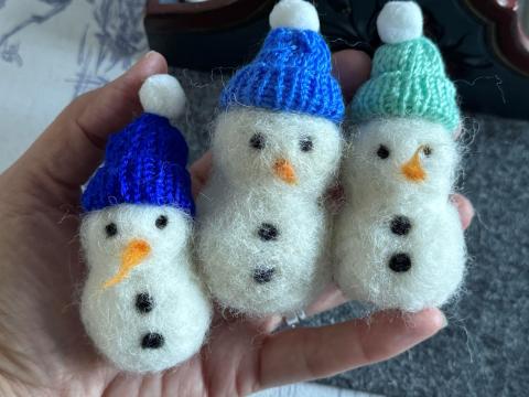 Photograph of three felted snowmen with knitted hats in various shades of blue held in the palm of someone's hand