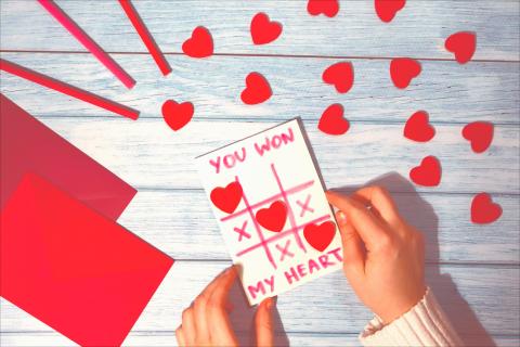 Photograph of two hands holding a small sheet of paper with tic-tac-toe board made up of hearts and X's, with text above reading "You won my heart!," a series of red pieces of paper (both full and cut into strands and hearts) laid atop a white wooden table