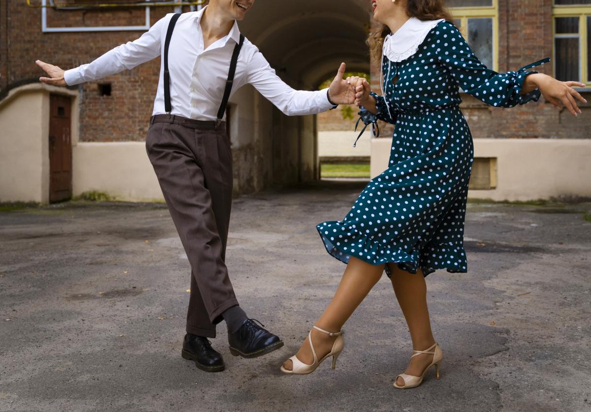 Man in white button down and dark trousers with suspenders dancing in a parking lot in a wide, upbeat manner with a woman in a turquoise dress with a white lapel and white polka dots